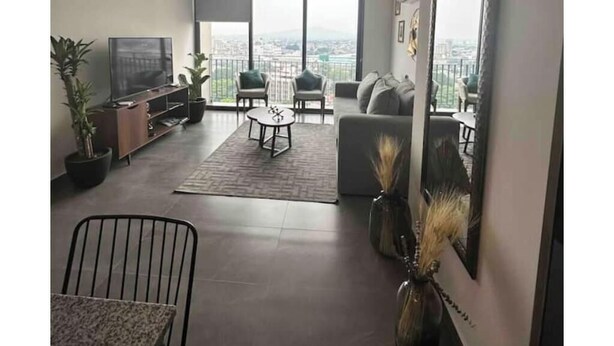 A modern living room with a TV, seating, a coffee table, and large windows offering a city view. - Cozy apartment in Americana 16 (Guadalajara)