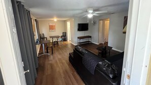En stue med en sort lædersofa, et spisebord med stole, en loftventilator og et fjernsyn monteret på væggen.