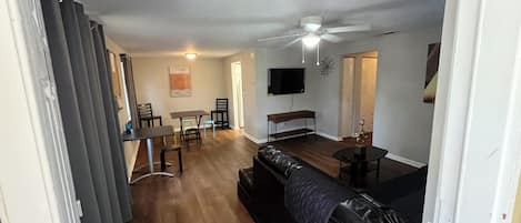 En stue med en sort lædersofa, et spisebord med stole, en loftventilator og et fjernsyn monteret på væggen.