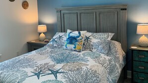 Un dormitorio con cama, ventilador de techo, mesita de noche con lámpara, cuadro enmarcado en la pared y almohadón decorativo.