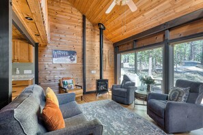 L’intérieur chaleureux d’un chalet avec une cheminée, des sièges confortables et de grandes fenêtres donnant sur la forêt.
