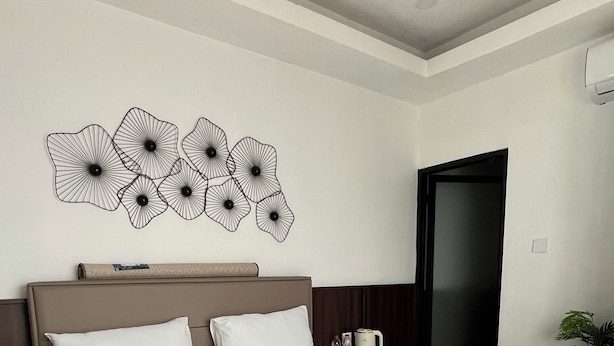 Ein Hotelzimmer mit einem großen Bett, einem Kopfteil, einer Wanddekoration, einem Nachttisch mit Tasse und Flasche sowie einer Tür.