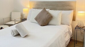Un lit bien fait, avec du linge de lit blanc et une serviette roulée, une tête de lit, des lampes de chevet et un tableau représentant un paysage marin.