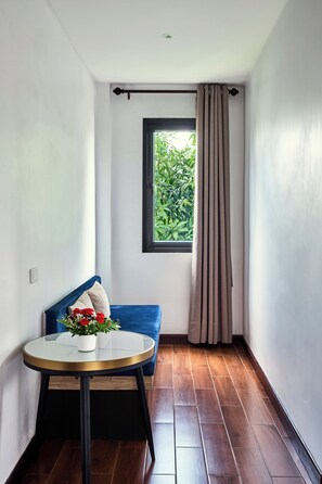 A room with a blue sofa, a round glass table with a flower vase, a window with a view of greenery, and a wooden floor. - RS1 Hotel (Vientiane)