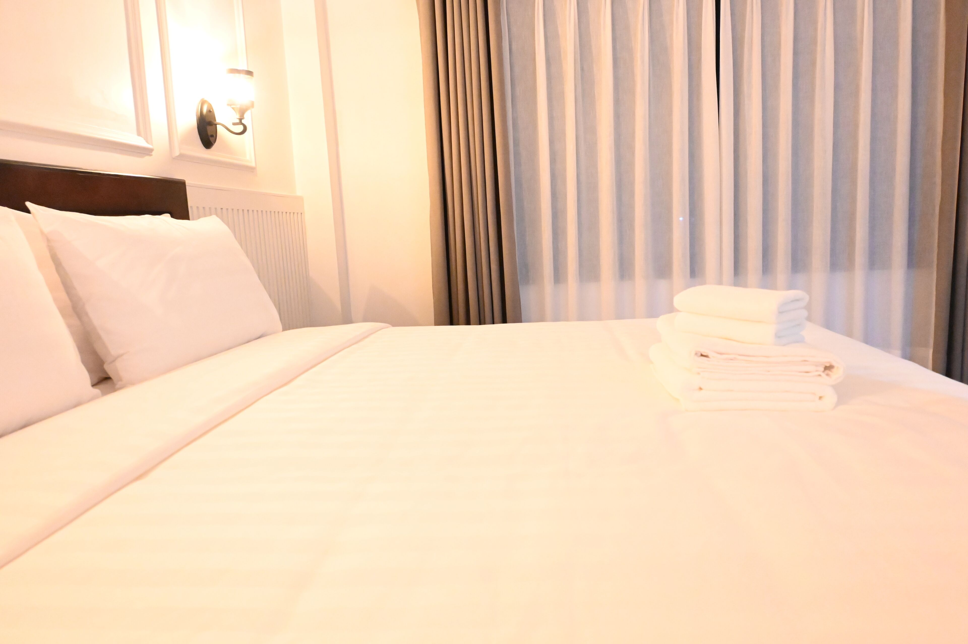 Una cama bien hecha con sábanas blancas, una pila de toallas dobladas y una lámpara de pared.