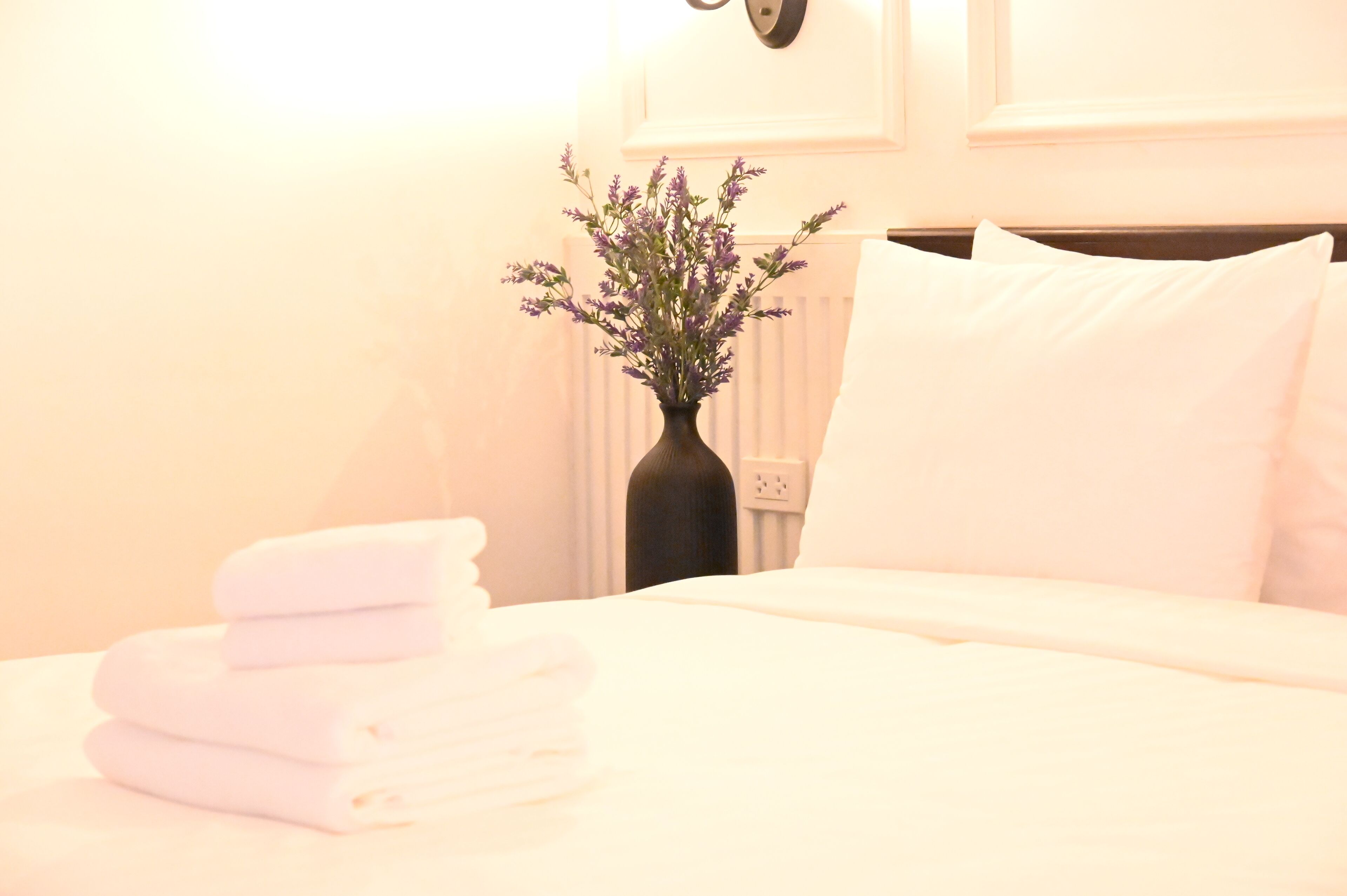 A neatly made bed with folded towels, a vase with flowers, and a wall-mounted light fixture.