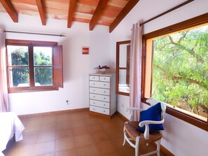A room with a wooden ceiling, a window with curtains, a white dresser, a wicker chair with a cushion, and a view of greenery outside. - Sa Taulera – Private Pool & WiFi in Capdepera (Capdepera)