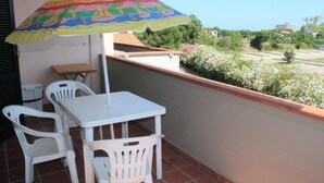 Un balcone con tavolo e sedie bianche, un ombrellone colorato e vista su spazi verdi ed edifici.