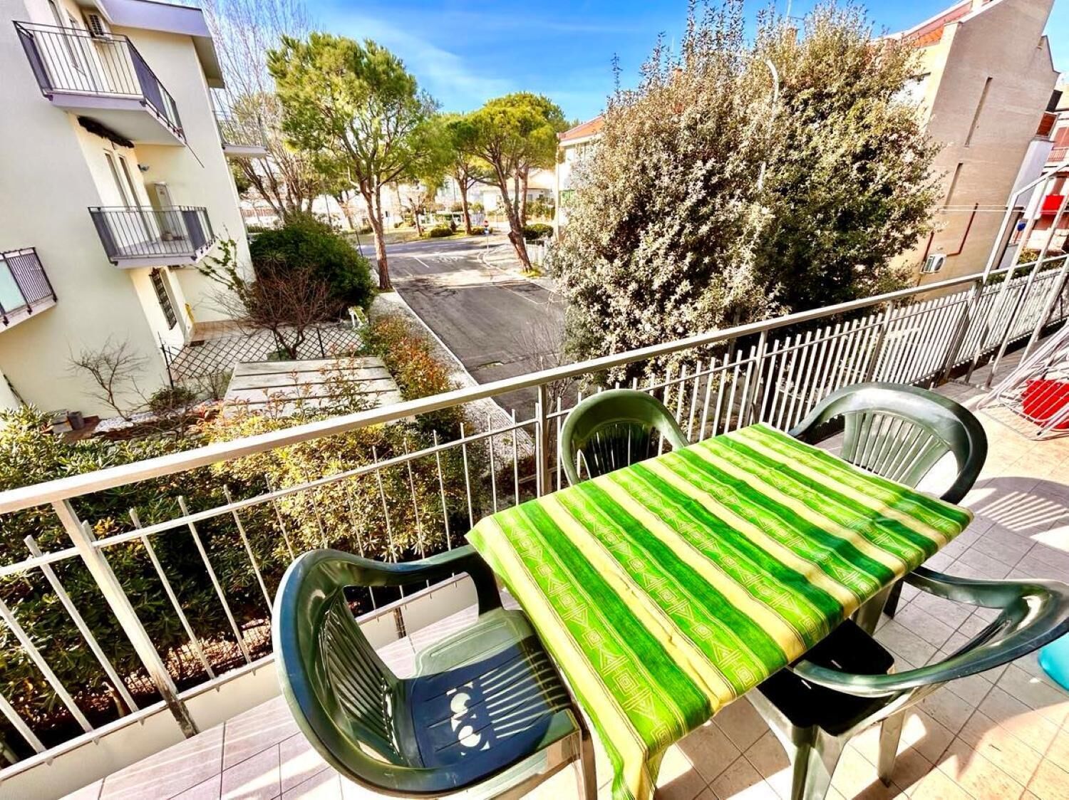 Un balcon avec une table et des chaises, donnant sur une rue bordée d’arbres et de bâtiments.