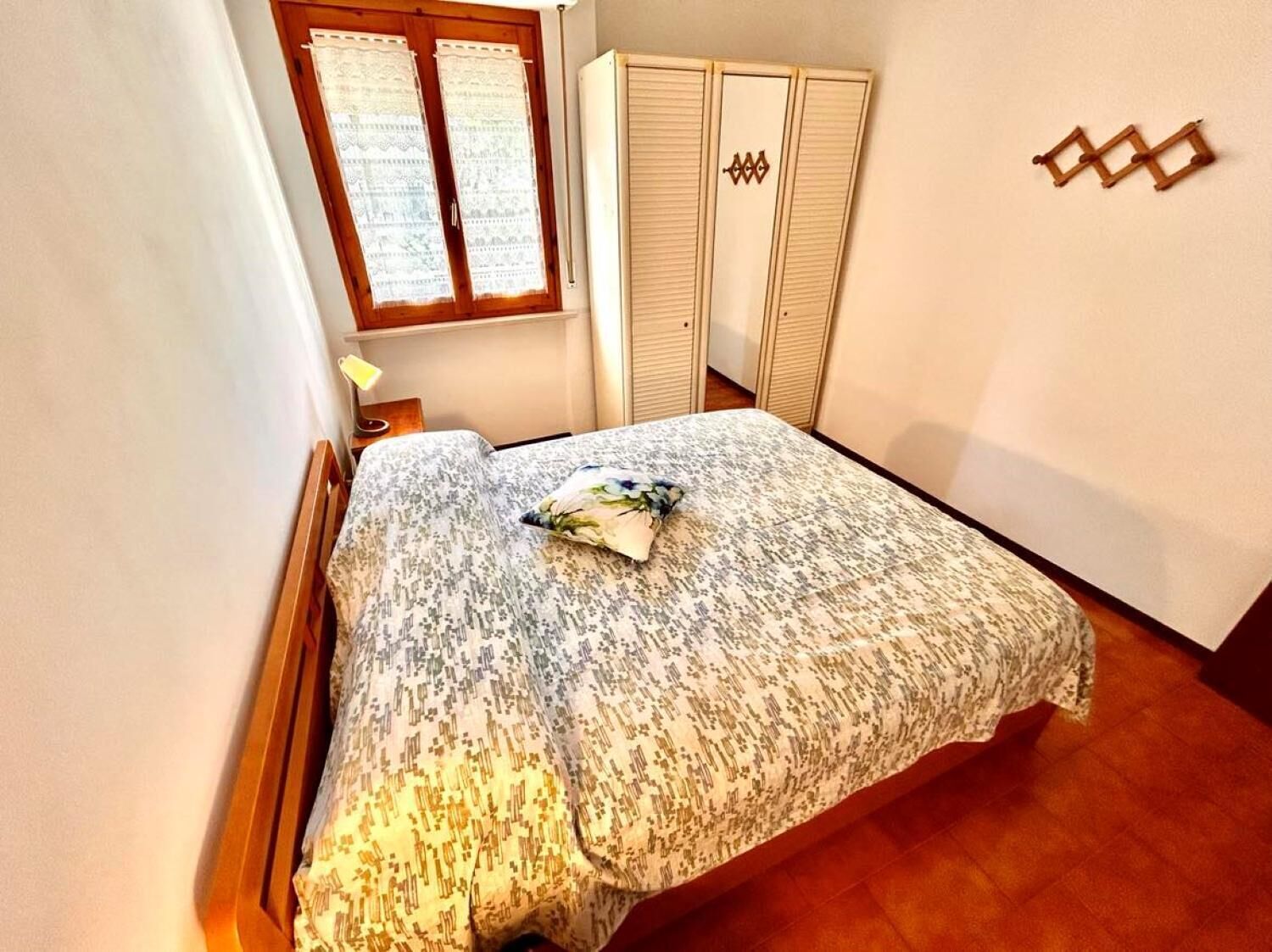 A bedroom with a bed, a window with lace curtains, a wardrobe, and a wall-mounted decoration.