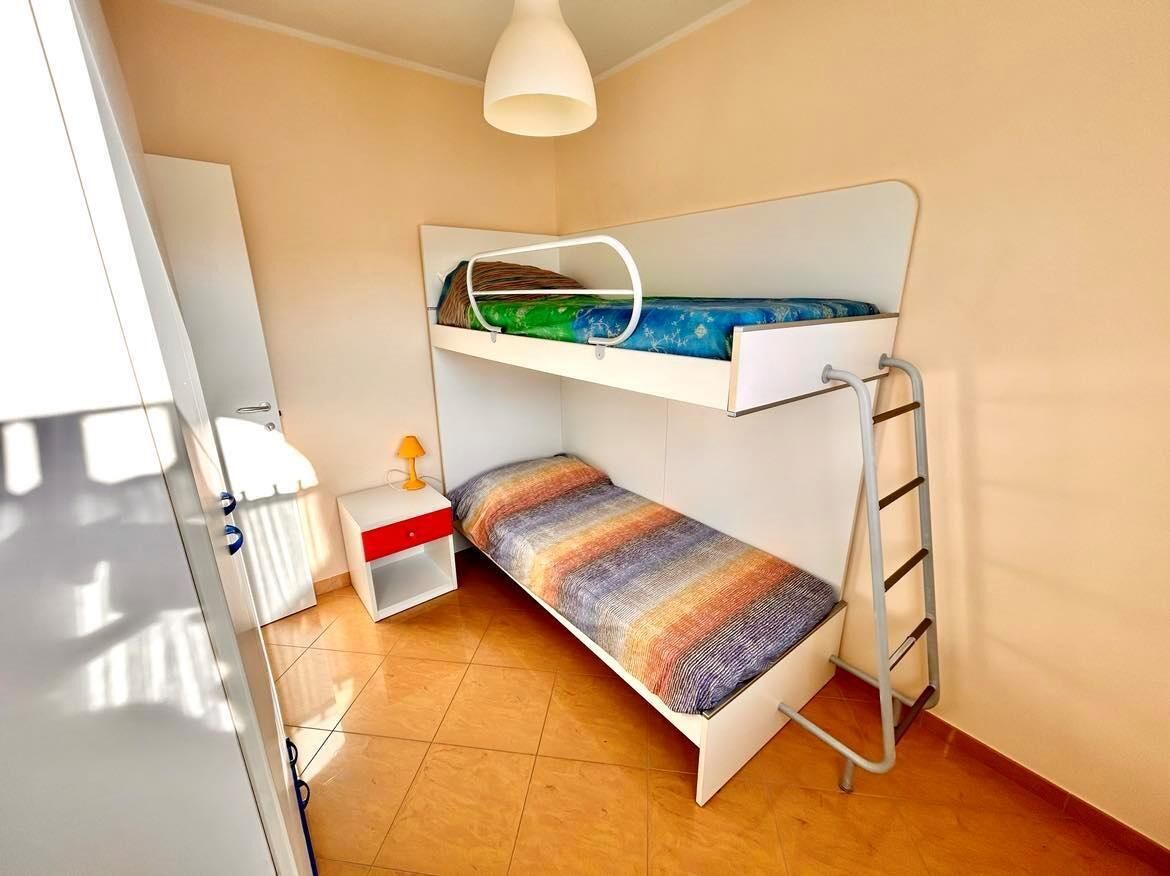 A small, compact room with a bunk bed, a small bedside table with a lamp, and a white wardrobe.