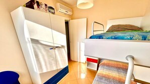 Ein kleines Zimmer mit Etagenbett, Schreibtisch, Stuhl, Kleiderschrank und einer Pendelleuchte.
