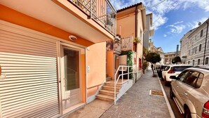 A narrow street with parked cars, a building with a closed shutter, and a staircase leading to a balcony. - Brand new one-bedroom apartment in downtown with 2 bathrooms and WI-FI (Numana)