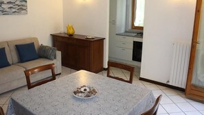 Een woonkamer met een bank, een eettafel met een schaal, een houten kast en een keuken zichtbaar door een boog.