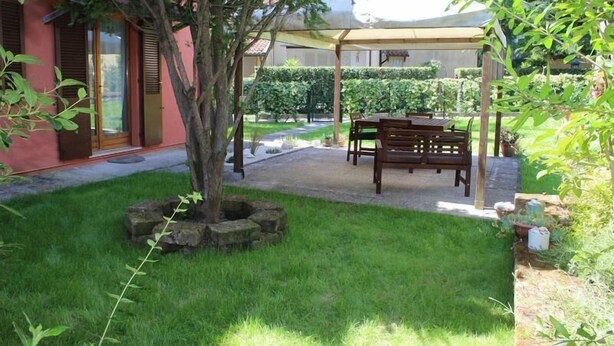 Een terras met een houten tafel en stoelen, een overdekte zithoek en een verzorgde tuin met een boom en stenen plantenbakken.