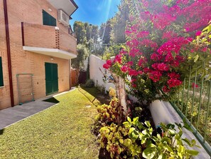 A residential area with a brick building, a small lawn, and flowering plants. - Cozy 1-bedroom apartment with a garden and parking near downtown (Sirolo)