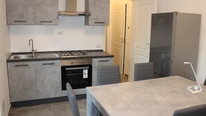 Ett kompakt kök med grå skåp, en diskho i rostfritt stål, en gasspis och en inbyggd ugn. Det finns ett matbord med en stol och en modern hänglampa.