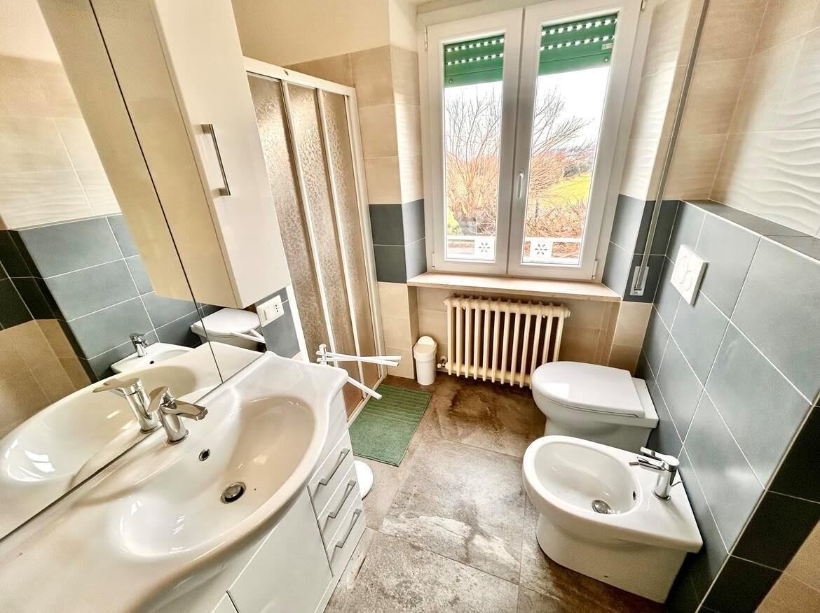 A bathroom with a double sink, toilet, bidet, and a window with a view of trees.