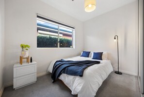 Une chambre à coucher avec un lit, une table de chevet ornée d’une plante, une fenêtre avec des stores et une lampe sur pied.