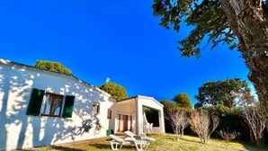 Uma casa branca com varanda coberta, um gramado com algumas cadeiras e árvores ao fundo.