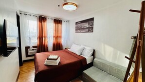 Quarto de hotel com cama, mesa de cabeceira, poltrona, televisão e janela com cortinas.