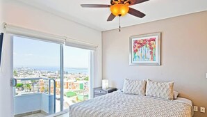 A bedroom with a bed, a ceiling fan, a window with a view, and a painting on the wall. - Oceanview Balcony, BBQ Lounge, Near Malecon (La Paz)