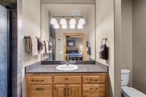 Un baño con encimera de granito, un lavabo blanco, armarios de madera y un espejo con luces arriba.