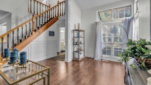 A spacious living area with a wooden staircase, a glass coffee table, and a large window with blinds. - Centrally Located Retreat (Indianapolis)