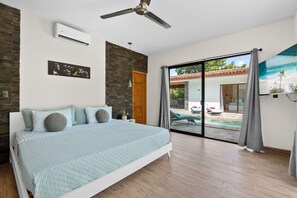 Un dormitorio con cama, ventilador de techo, equipo de aire acondicionado, puerta de madera y vista a una piscina exterior.