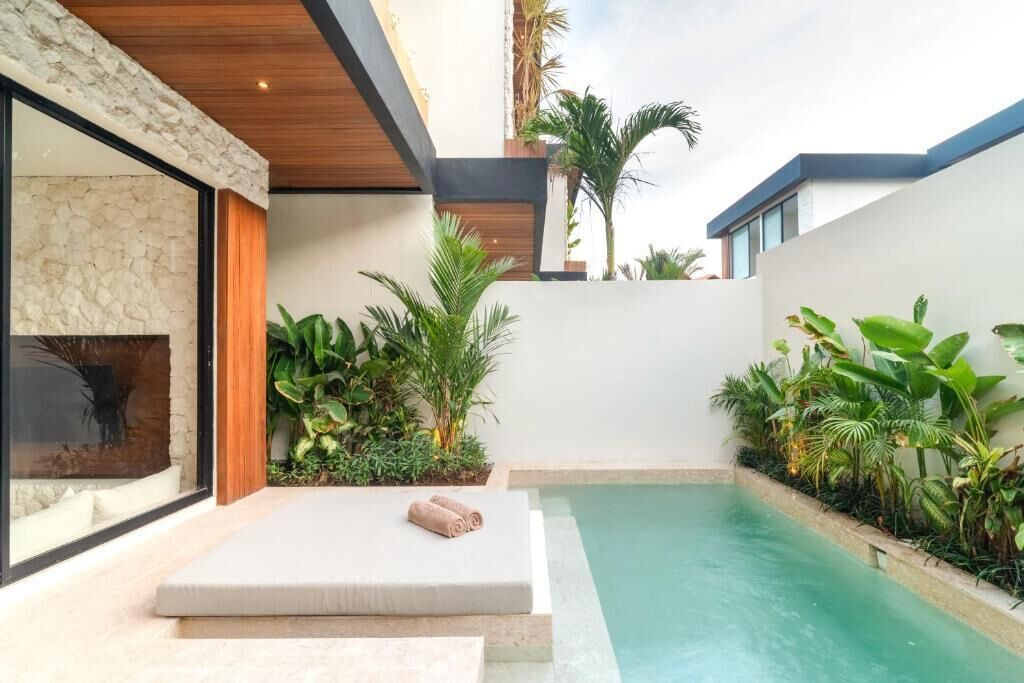 Un'area piscina moderna con una poltrona da relax e cuscini, immersa nel verde e delimitata da un muro in pietra.