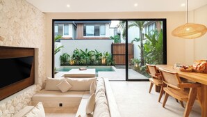 A modern living room with a stone wall, a flat-screen TV, a sofa, a dining area with wooden furniture, and a view of a pool and greenery outside. - La Finca Villa Bingin, Uluwatu by Sandara (Pecatu)