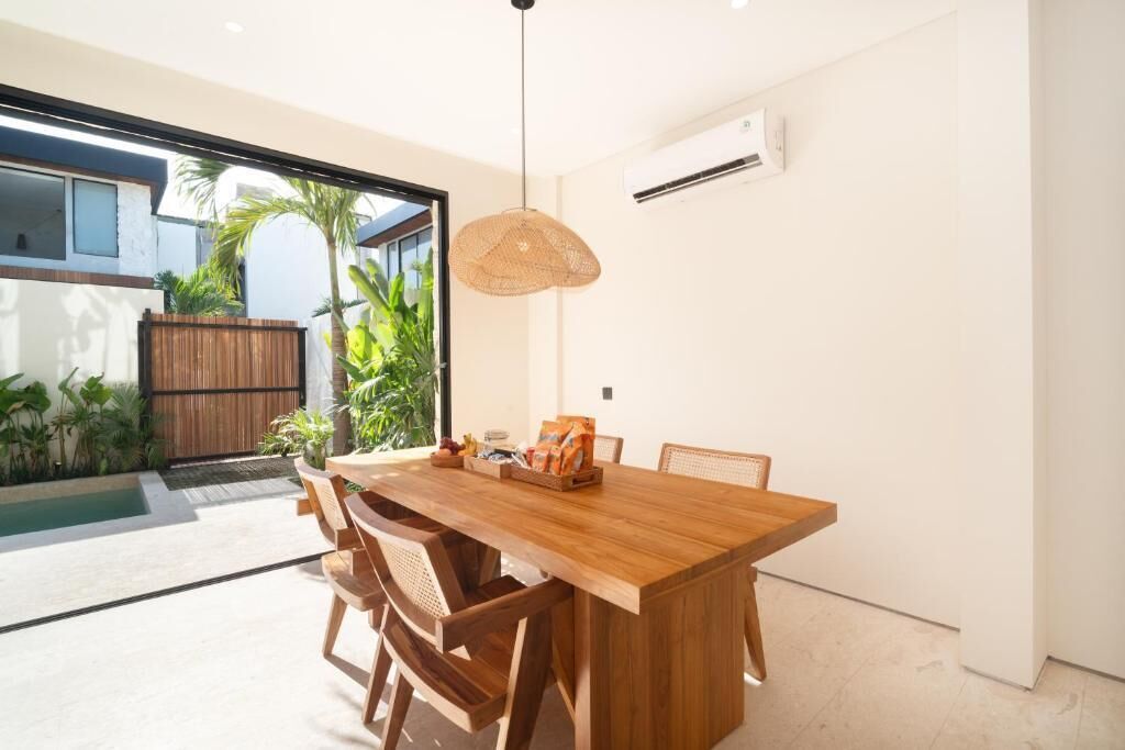 A modern dining area with a wooden table and chairs, a hanging light fixture, and a view of a pool and greenery outside.