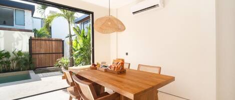 A modern dining area with a wooden table and chairs, a hanging light fixture, and a view of a pool and greenery outside.