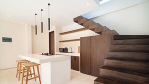 A modern kitchen with a central island, wooden stools, a built-in microwave, and a staircase with wooden steps. - La Finca Villa Bingin, Uluwatu by Sandara (Pecatu)