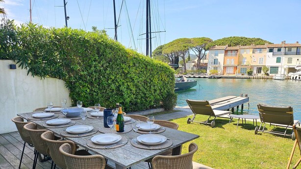 Un espace de restauration en plein air avec une table dressée pour six personnes, entouré de chaises en osier, offrant une vue sur une étendue d’eau où l’on aperçoit des bateaux et des bâtiments au loin.