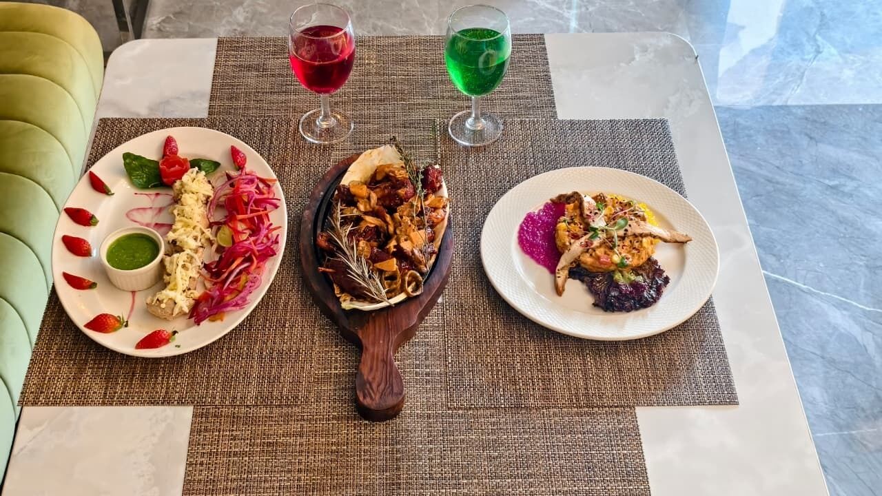 Een tafel met drie borden eten, twee glazen gekleurde drankjes en een houten serveerplank.