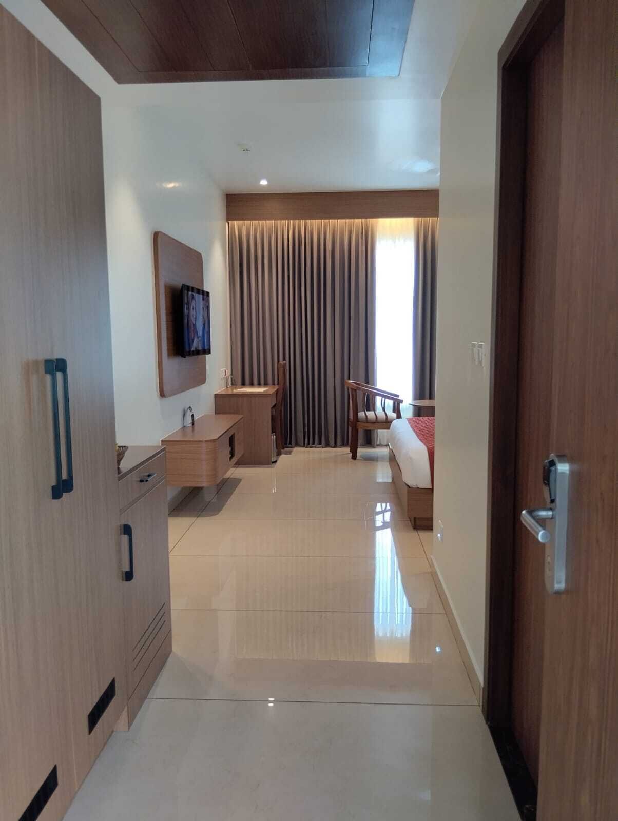 Habitación de hotel moderna con armario de madera, escritorio, silla, cama, televisor montado en la pared y un amplio ventanal con cortinas.