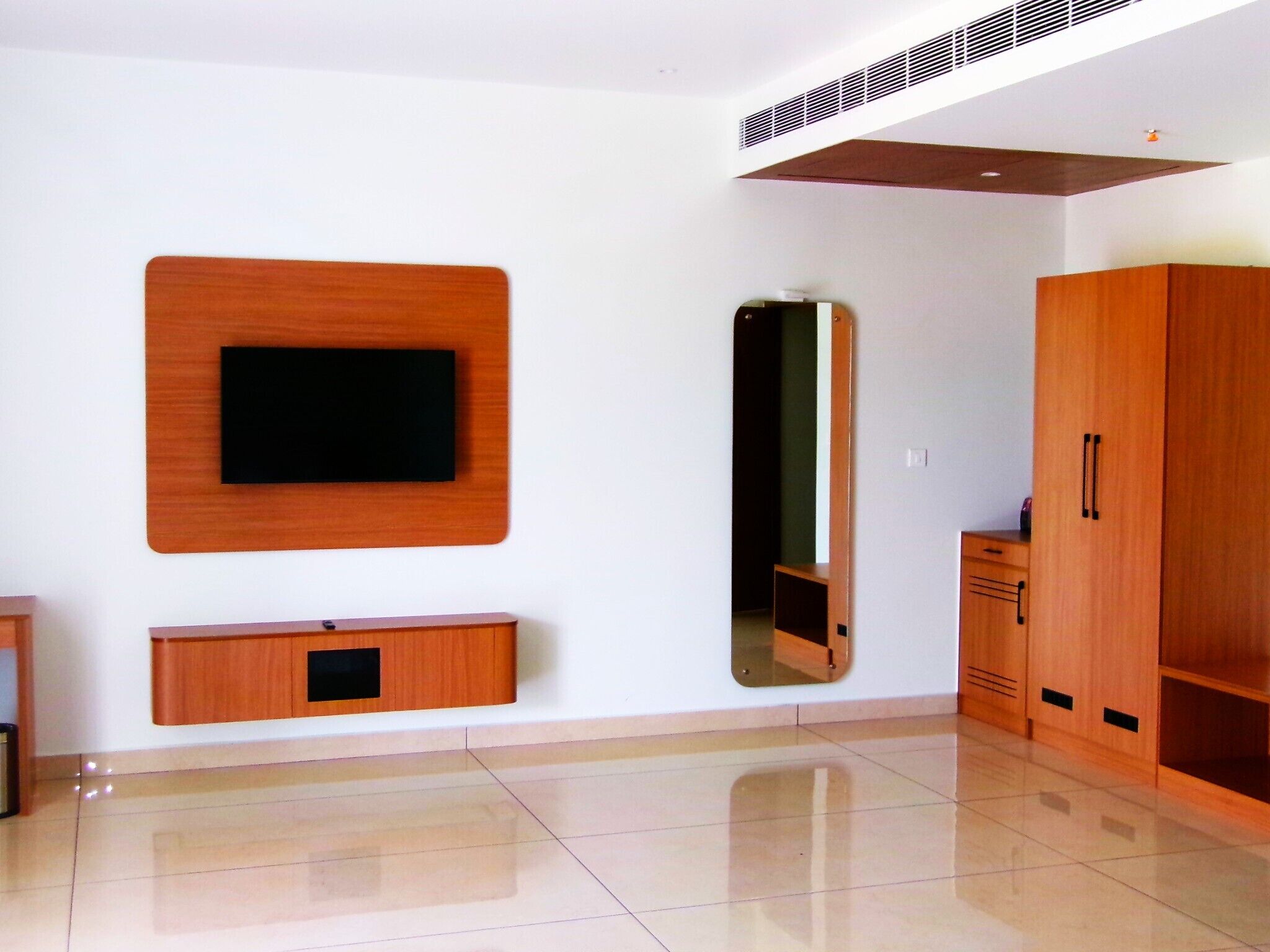 A modern room with a flat-screen TV mounted on the wall, a large mirror, wooden cabinets, and a tiled floor.