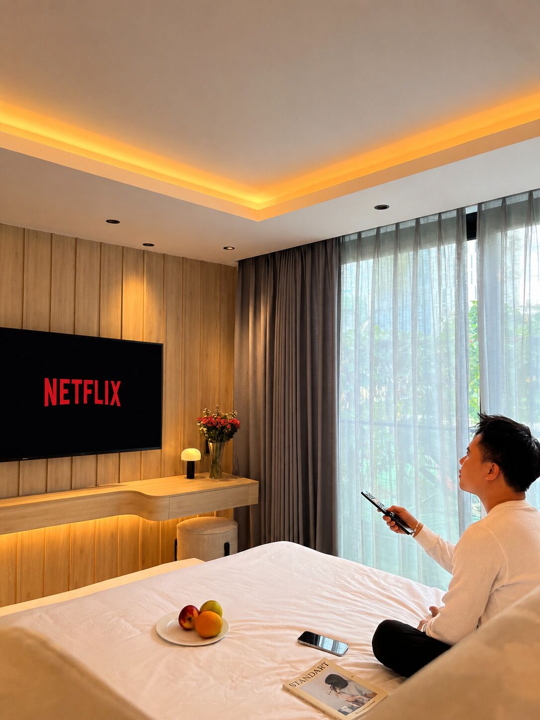 Et hotelværelse med en seng, et fjernsyn der viser 'NETFLIX', et natbord med en lampe og blomster, samt et vindue med gardiner.