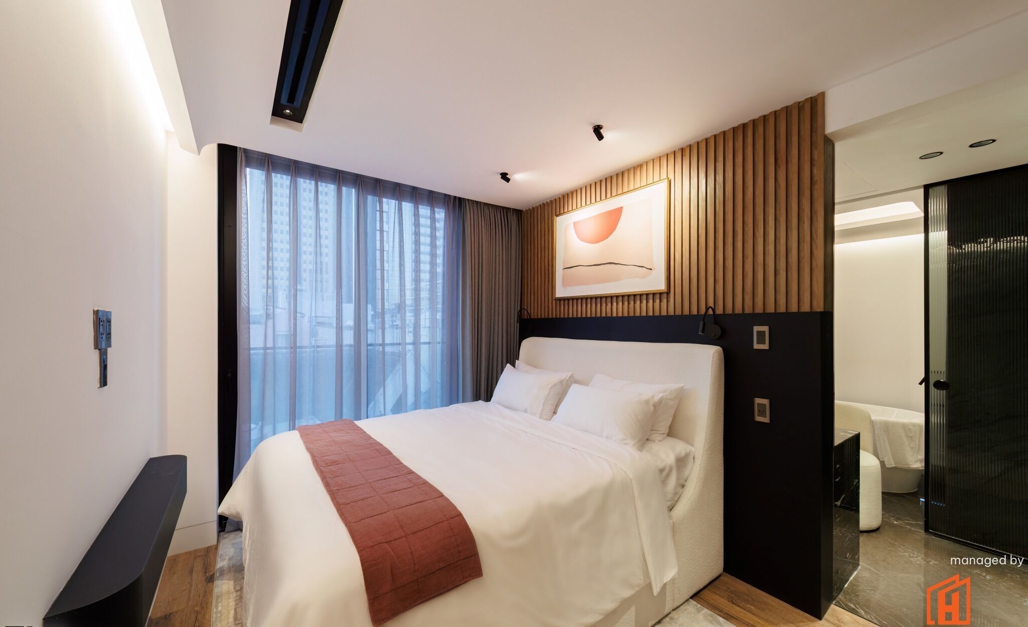 A modern hotel room with a large bed, a wooden panel wall, a painting, and a bathroom with a glass door.