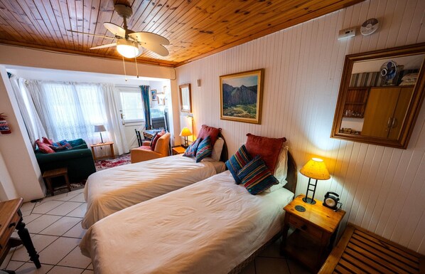 Une chambre avec deux lits, un ventilateur de plafond, une fenêtre avec des rideaux, un tableau au mur et un miroir.