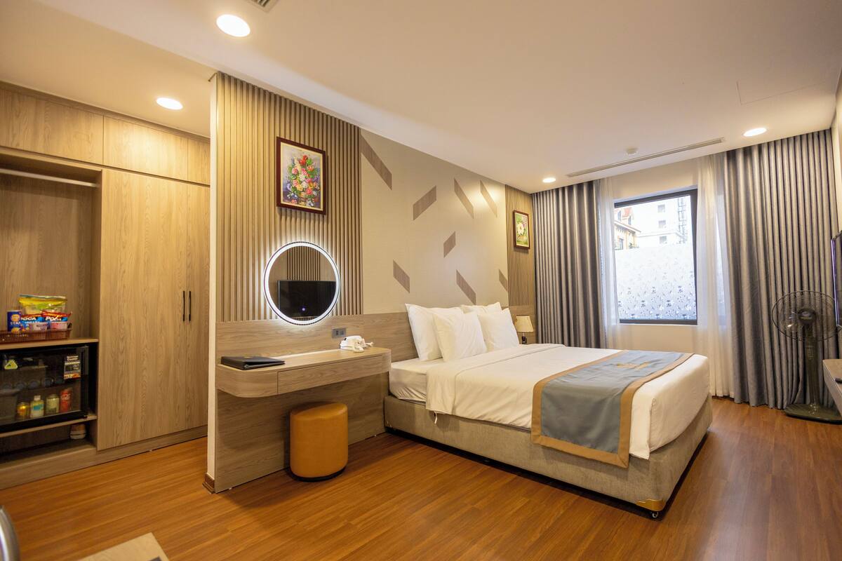 A hotel room with a bed, a desk, a vanity with a mirror, a wardrobe, and a window with curtains.