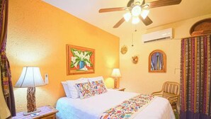 Une chambre d’hôtel comprenant un lit, deux lampes de chevet, un ventilateur de plafond, un appareil de climatisation, une chaise et un tableau décoratif au mur.