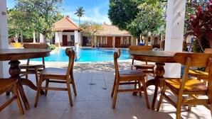 A poolside area with wooden tables and chairs, a clear blue pool, and a building with a tiled roof in the background. - Grand Melka Hotel (Buleleng)