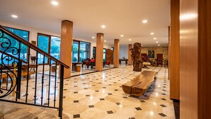A spacious lobby with a marble floor, wooden columns, a staircase with wrought iron railings, and large windows offering a view of the outdoors. - Grand Melka Hotel (Buleleng)