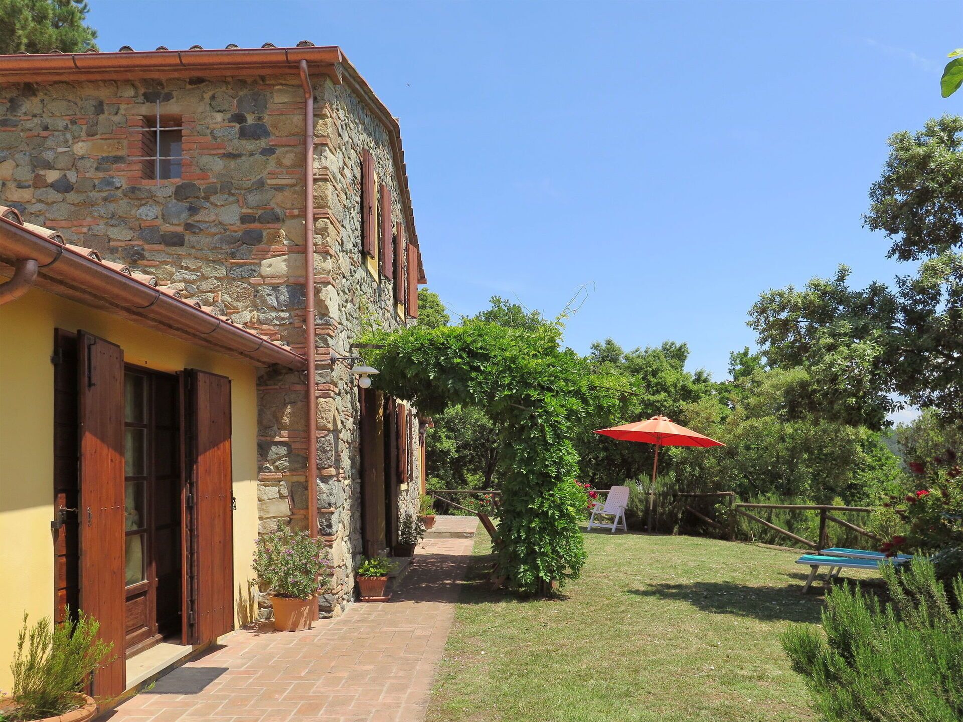 A stone building with a tiled roof, a paved pathway, and a garden with a red umbrella.