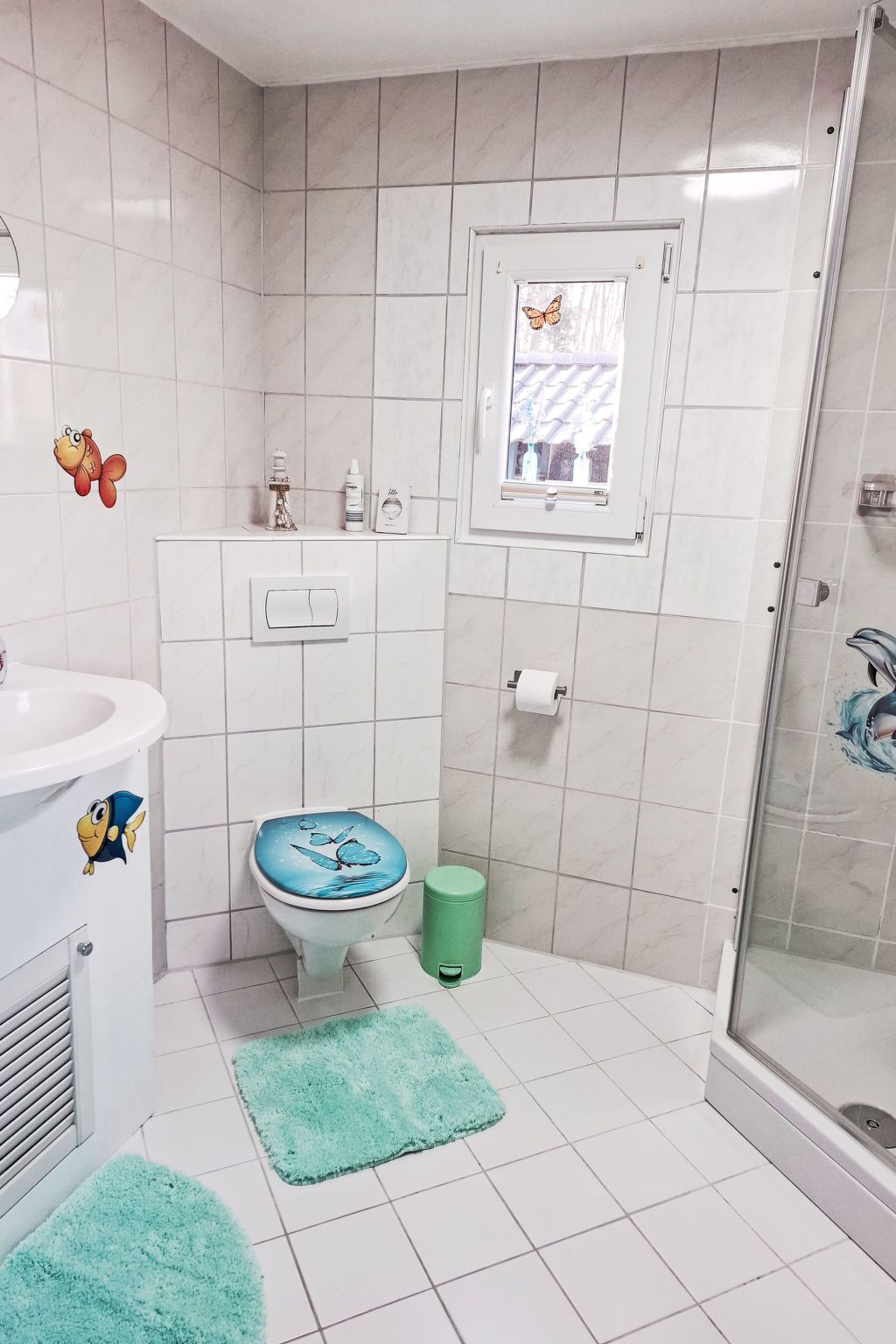 A bathroom with a toilet, sink, shower, and a window with a butterfly decal.