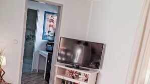 A living room with a flat-screen TV on a white stand, a glass coffee table with magazines, a grey sofa, and a small grey ottoman. - Ferienbungalow auf Gartengrundstück by Interhome (Bad Doberan)