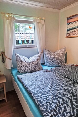 Ein Schlafzimmer mit Bett, Kissen, einem Fenster mit Vorhängen, einem Gemälde an der Wand und einem kleinen Hocker.