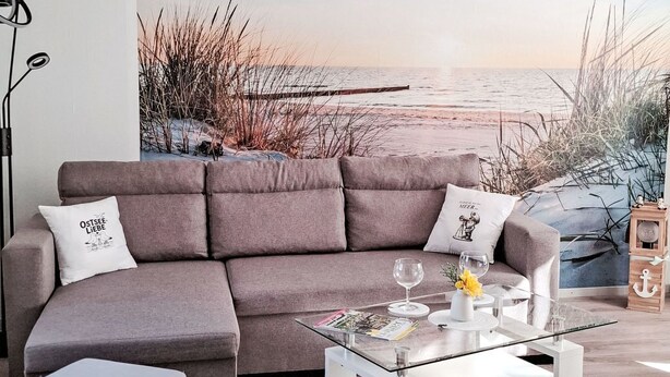 A modern living room with a sectional sofa, a glass coffee table, and a large beach-themed wall mural. - Ferienbungalow auf Gartengrundstück by Interhome (Bad Doberan)